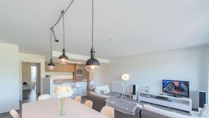 Una sala moderna con comedor, una cocina al fondo y un televisor sobre un mueble blanco.