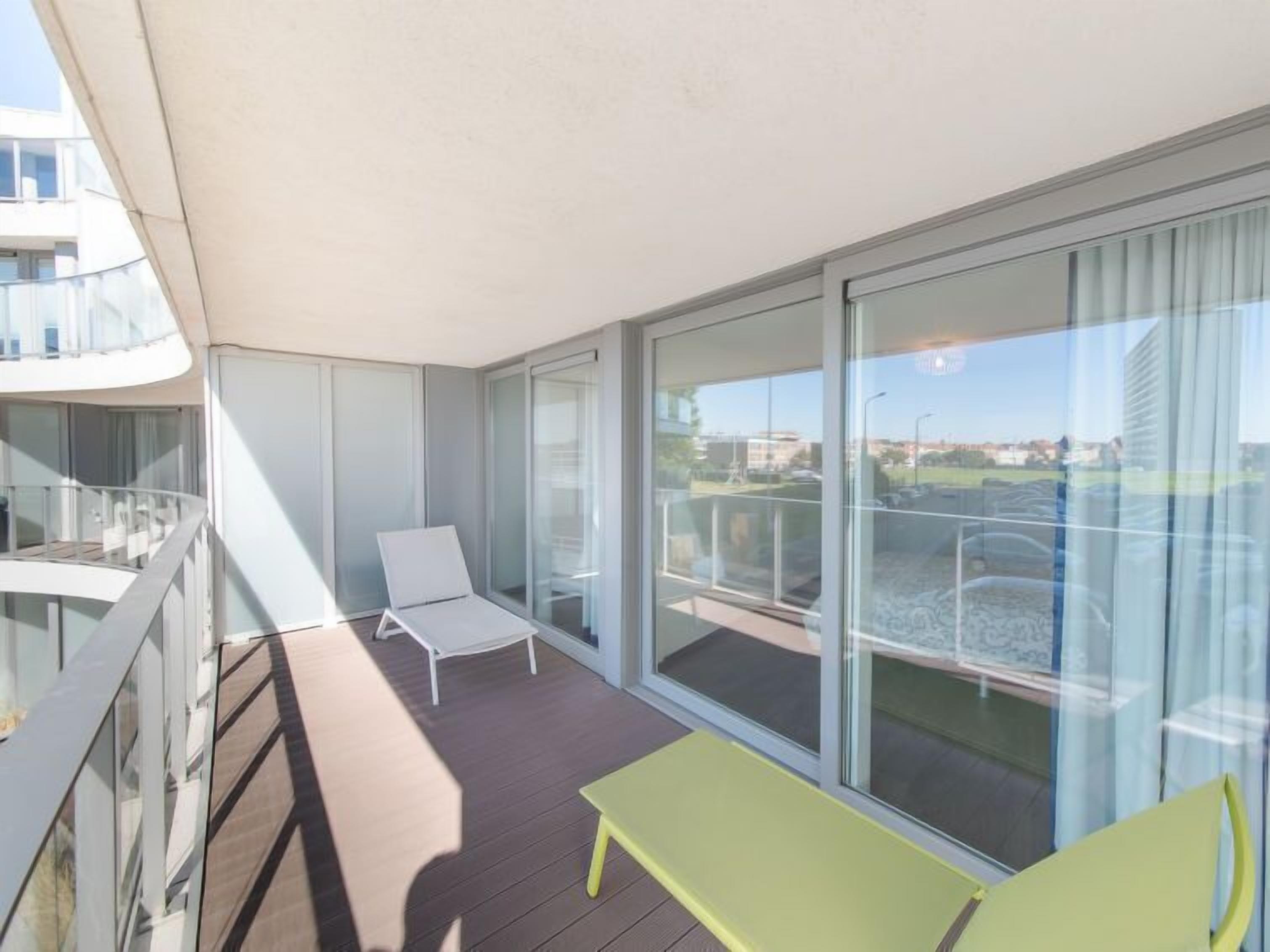 A balcony with a white lounge chair, a yellow table, and a glass door leading to a view of buildings and a road.