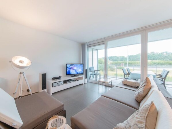 A modern living room with a flat-screen TV, a white entertainment unit, and a large window offering a view of greenery.