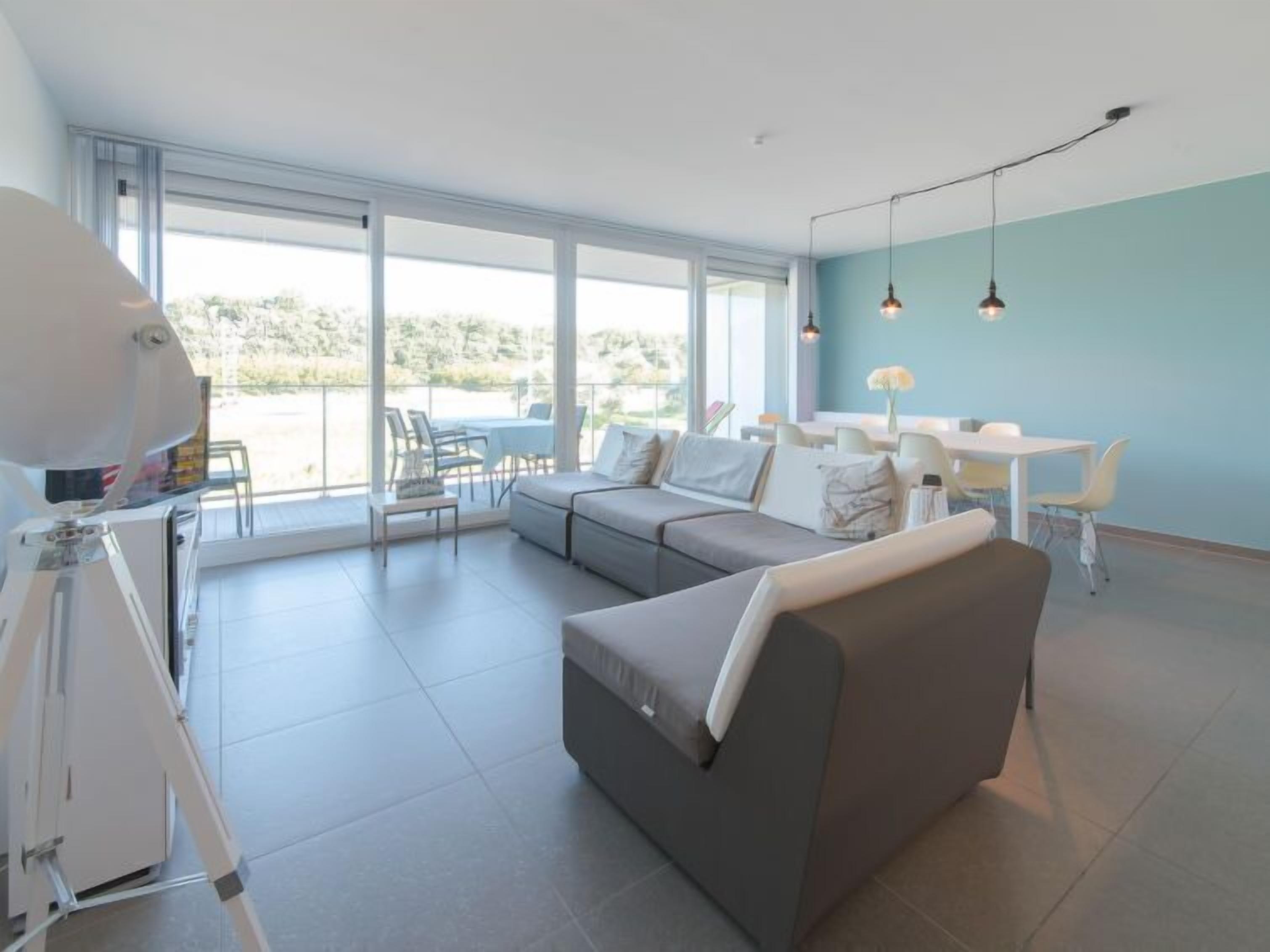 Una sala moderna con un sofá gris, un comedor con sillas blancas y ventanas grandes que ofrecen vistas al exterior.