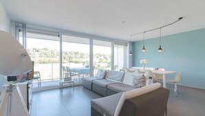 Una sala moderna con un sofá gris, un comedor con sillas blancas y ventanas grandes que ofrecen vistas al exterior.