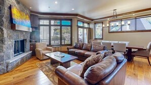 A spacious living room with a stone fireplace, leather sofas, and large windows offering views of the outside. - Luxury Ski in, Ski out 3 Bedroom Mountain Residence in the Heart of Lionshead Village with Heated Pool and Slopeside Hot Tub (Vail)
