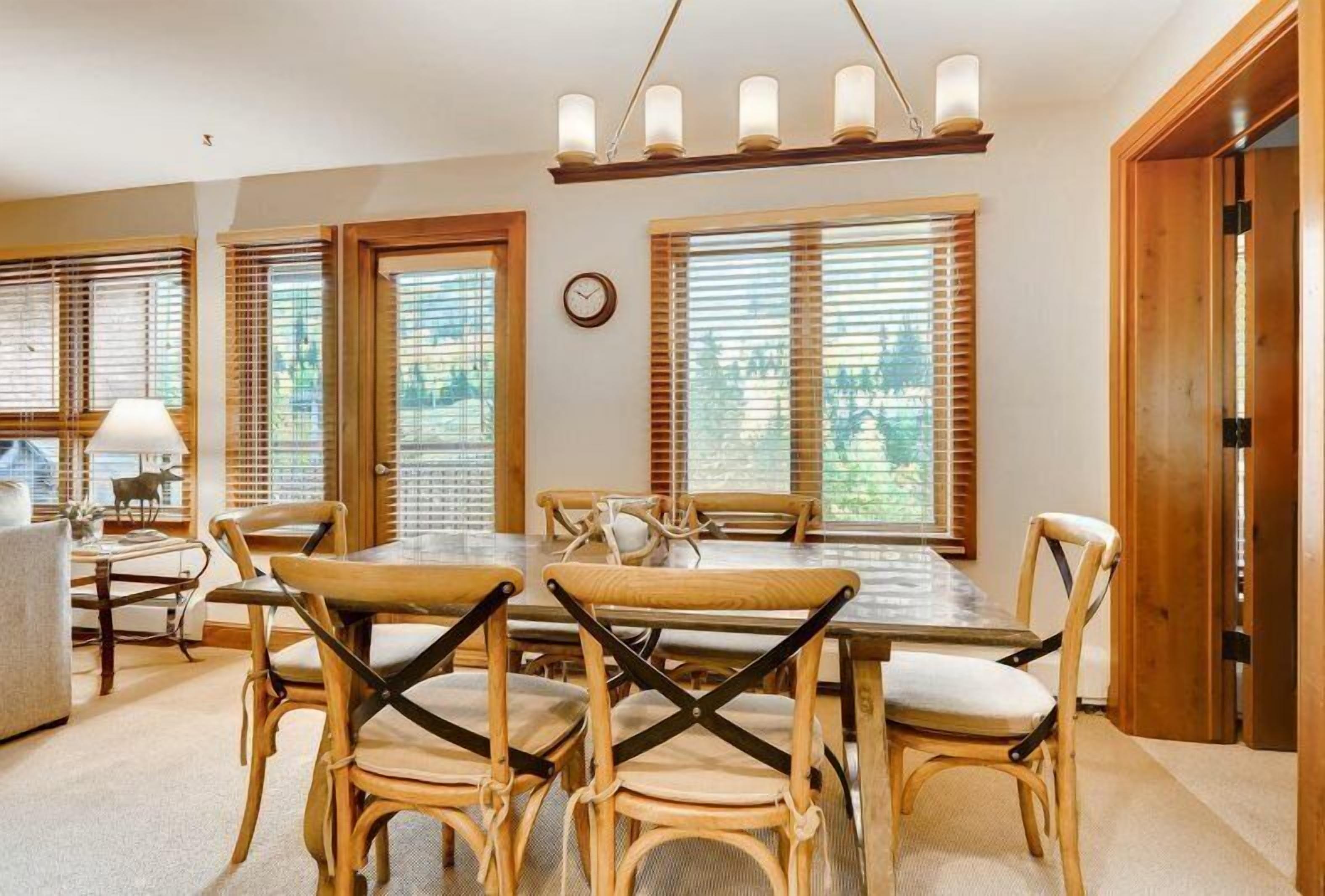 A dining area with a wooden table and chairs, large windows with blinds, and a chandelier.