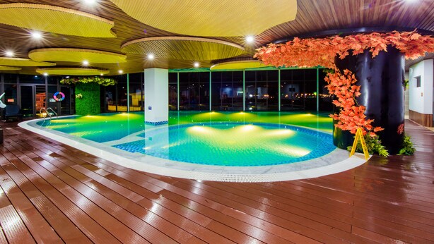 A swimming pool with a wooden deck, surrounded by a modern ceiling with circular patterns and a decorative plant with red leaves. - Sakura Hotel (Hoa Binh)