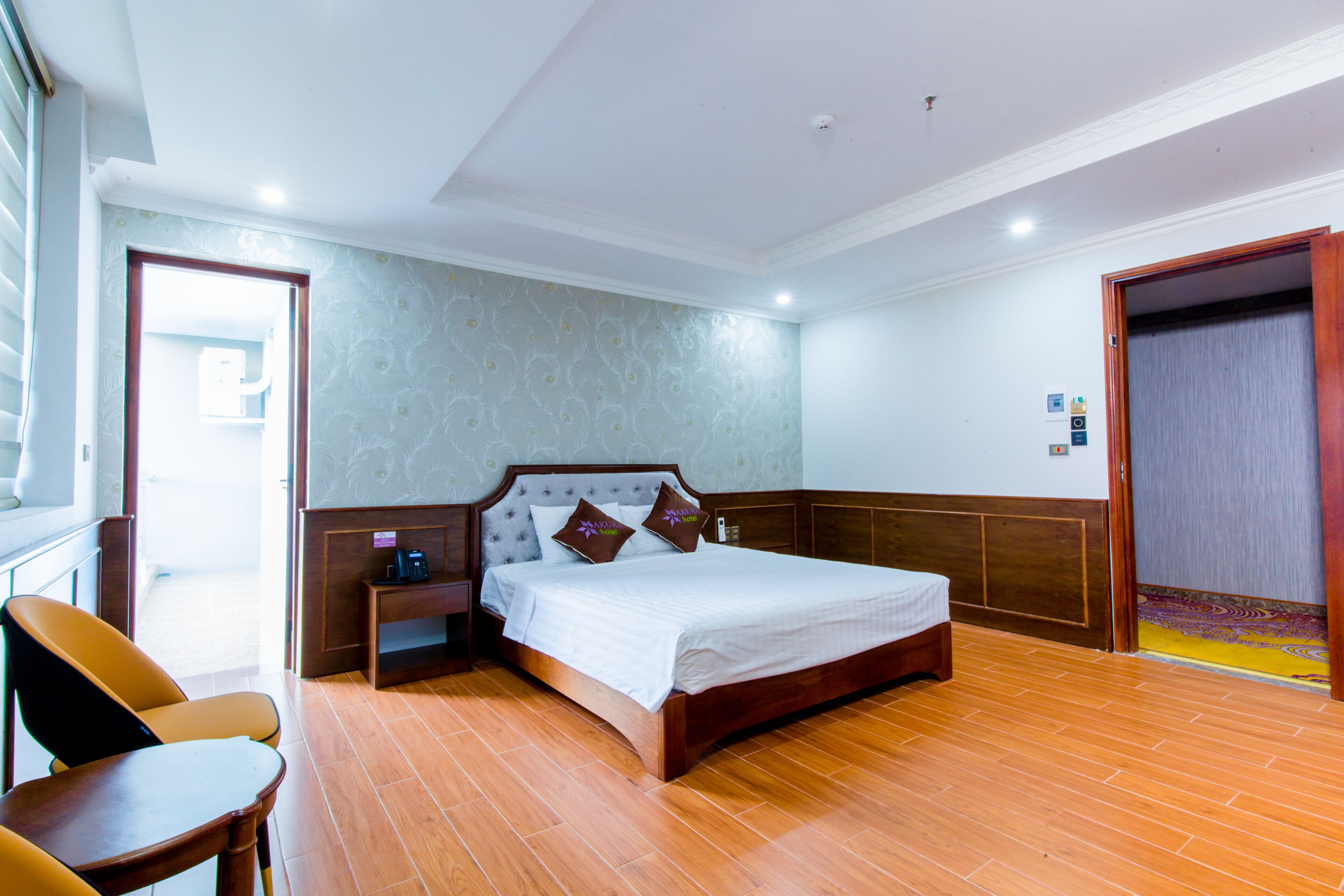 Quarto de hotel com uma cama grande, cabeceira de madeira e uma mesa de cabeceira com abajur. Há uma cadeira e uma pequena mesa no canto.