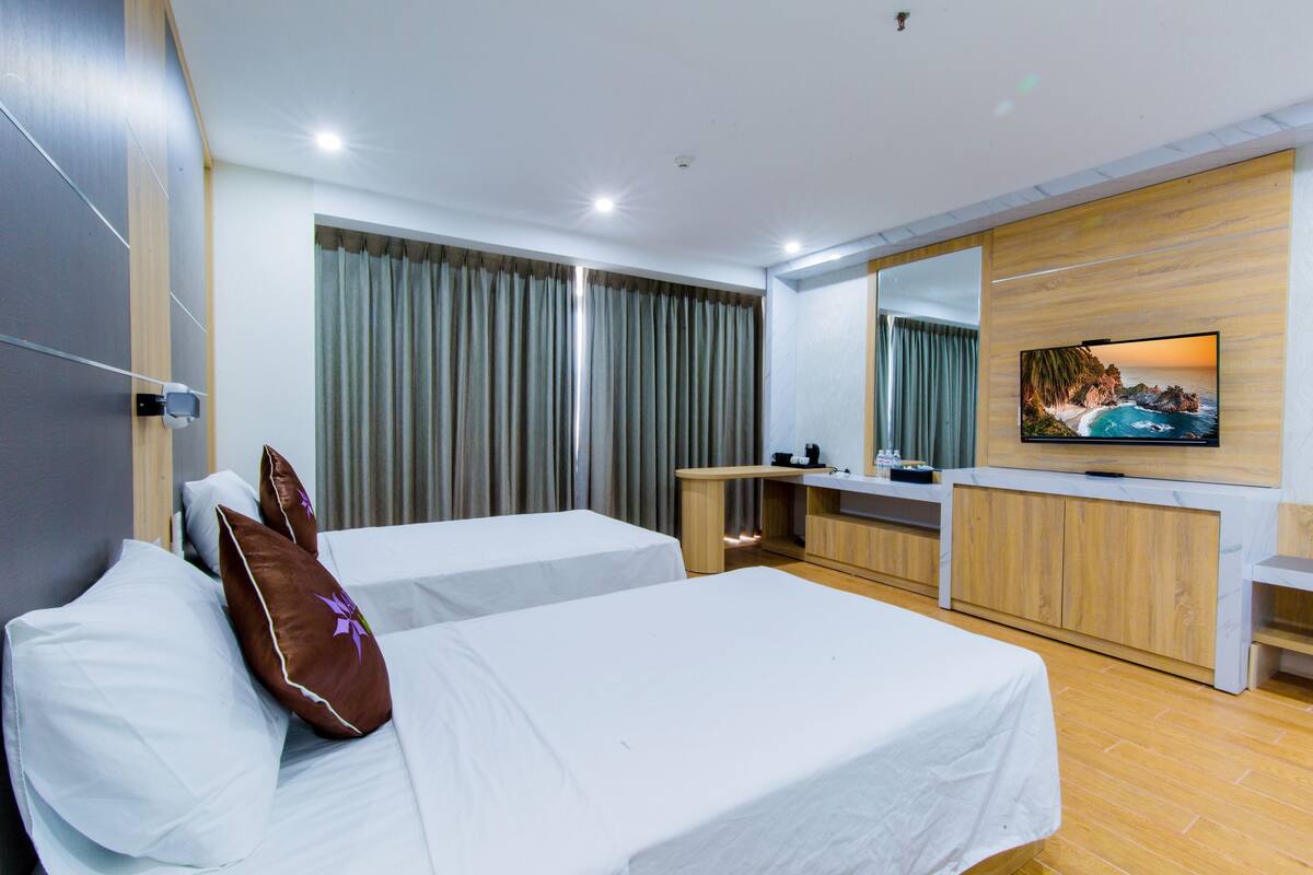 A hotel room with two beds, a wooden desk, a TV, and a large window with curtains.