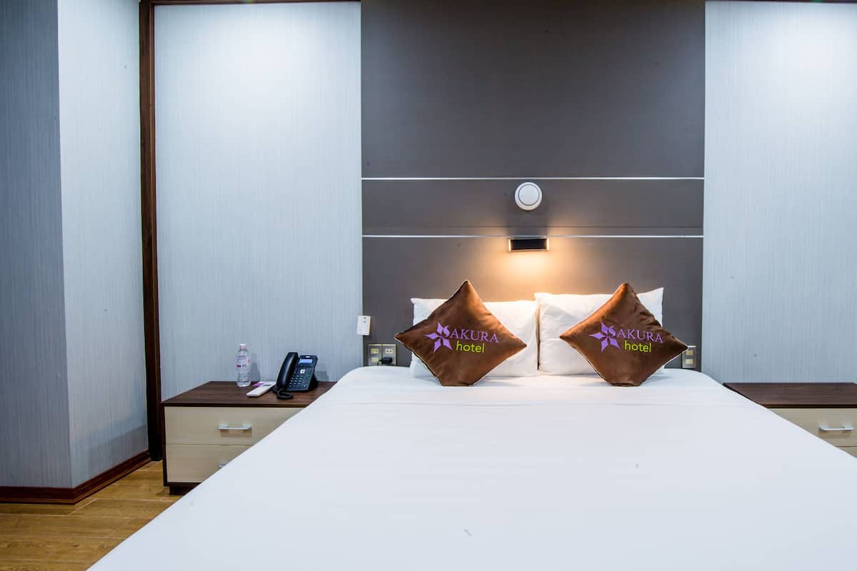 A hotel room with a large bed, two pillows with the hotel's logo, a bedside phone, a water bottle, and a clock on the wall.