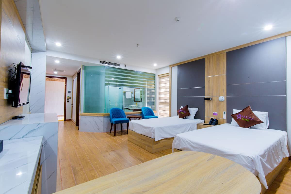 A hotel room with two beds, a desk, a chair, a small table, and a bathroom visible through a glass partition.