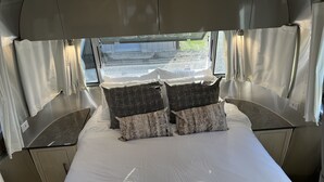 Una cama con ropa de cama blanca y dos almohadas oscuras con diseños, una ventana con cortinas, y vistas al exterior.