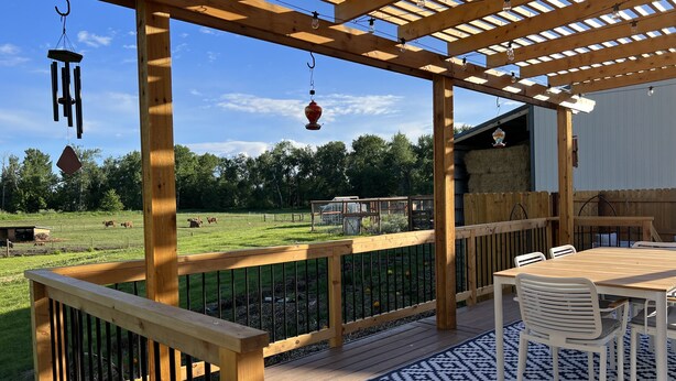 Una terraza de madera con un pérgola, mobiliario exterior y vistas a un pastizal con vacas.