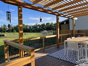 Eine Holzterrasse mit einer Pergola, Sitzgelegenheiten im Freien und Blick auf eine Weide mit Kühen.