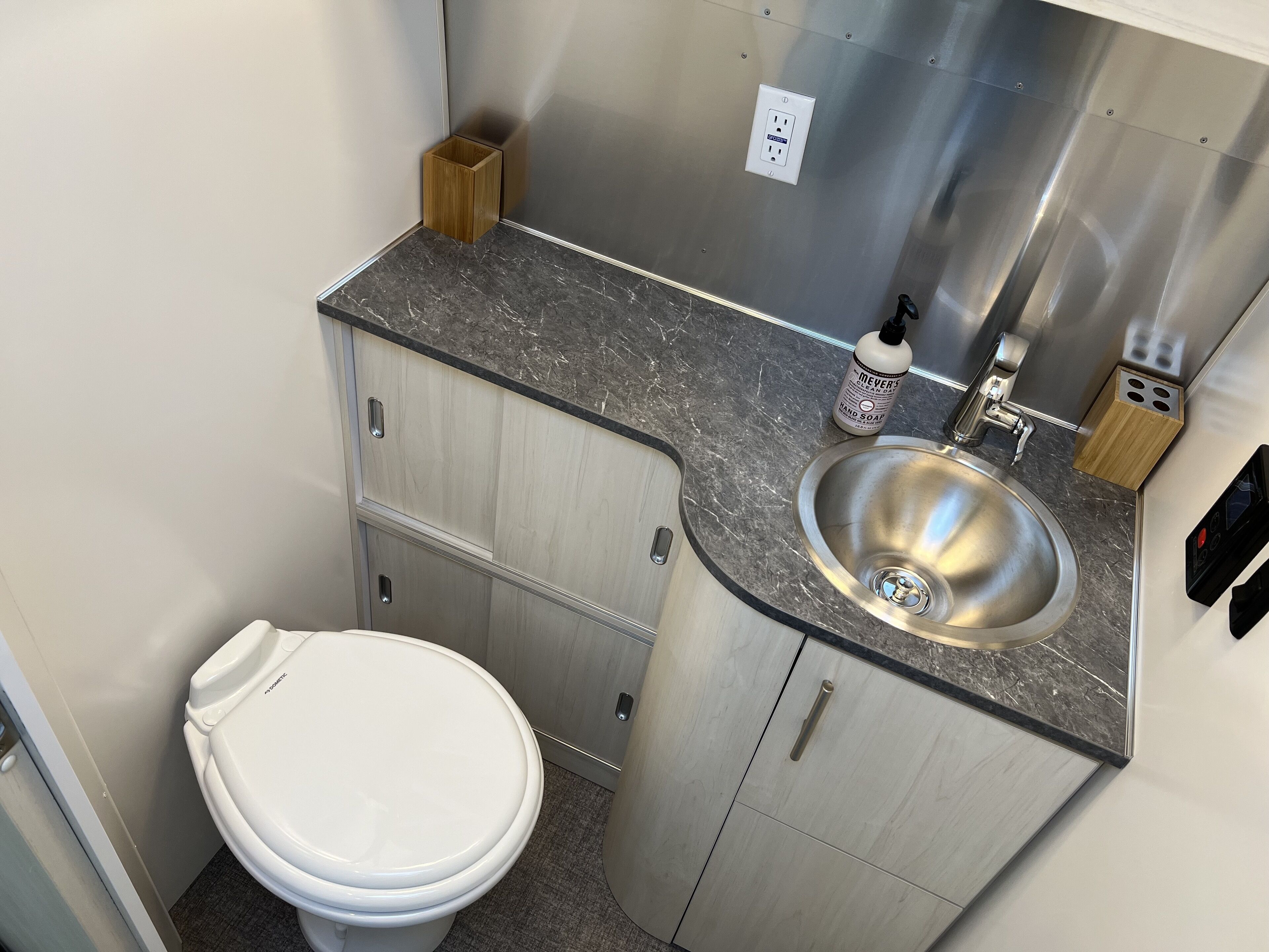 A compact bathroom with a white toilet, a stainless steel sink, and a wooden soap dispenser.