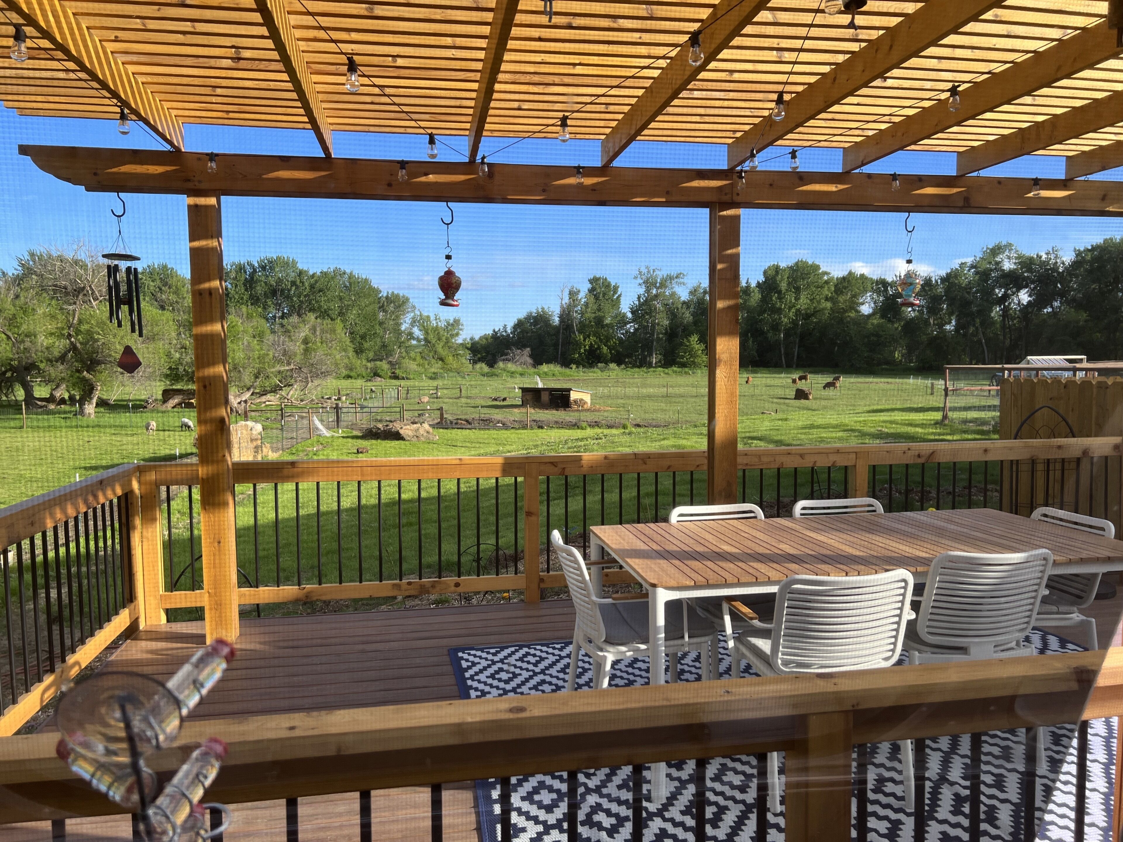 A wooden deck with a table and chairs, overlooking a grassy area with trees and a fenced enclosure.