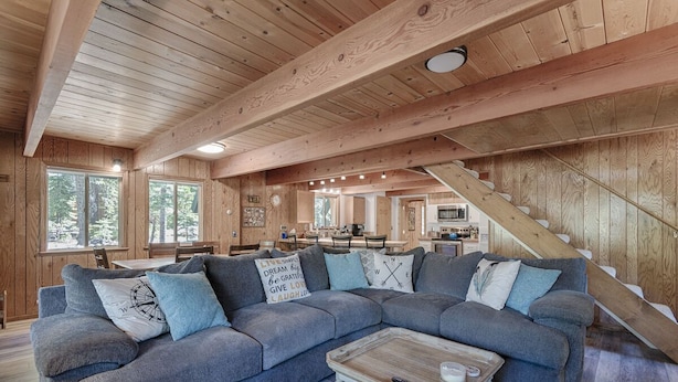 A cozy cabin interior with a large gray sectional sofa, wooden beams, a staircase, and a kitchen area. - Close drive to lake * Dog OK * Great Family Home (Carnelian Bay)