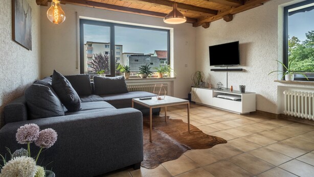 Uma sala de estar moderna com um sofá cinzento, uma mesa de centro, uma TV na parede e uma grande janela com vista para edifícios e árvores.