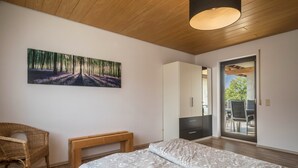 Um quarto com uma cama, um banco, uma cadeira de vime, um guarda-roupa e um quadro de uma floresta.