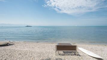 Ein Kiesstrand mit einer Holzbank und einem Surfbrett, ein Boot in der Ferne und ein klarer Himmel.