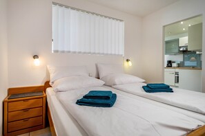 A double bed with white linens and two blue towels, a wooden nightstand with a drawer, a wall-mounted light, and a window with blinds.
