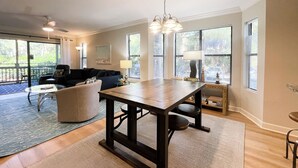 A spacious living room with a large wooden table, a dark-colored sofa, and a balcony with a view of trees.