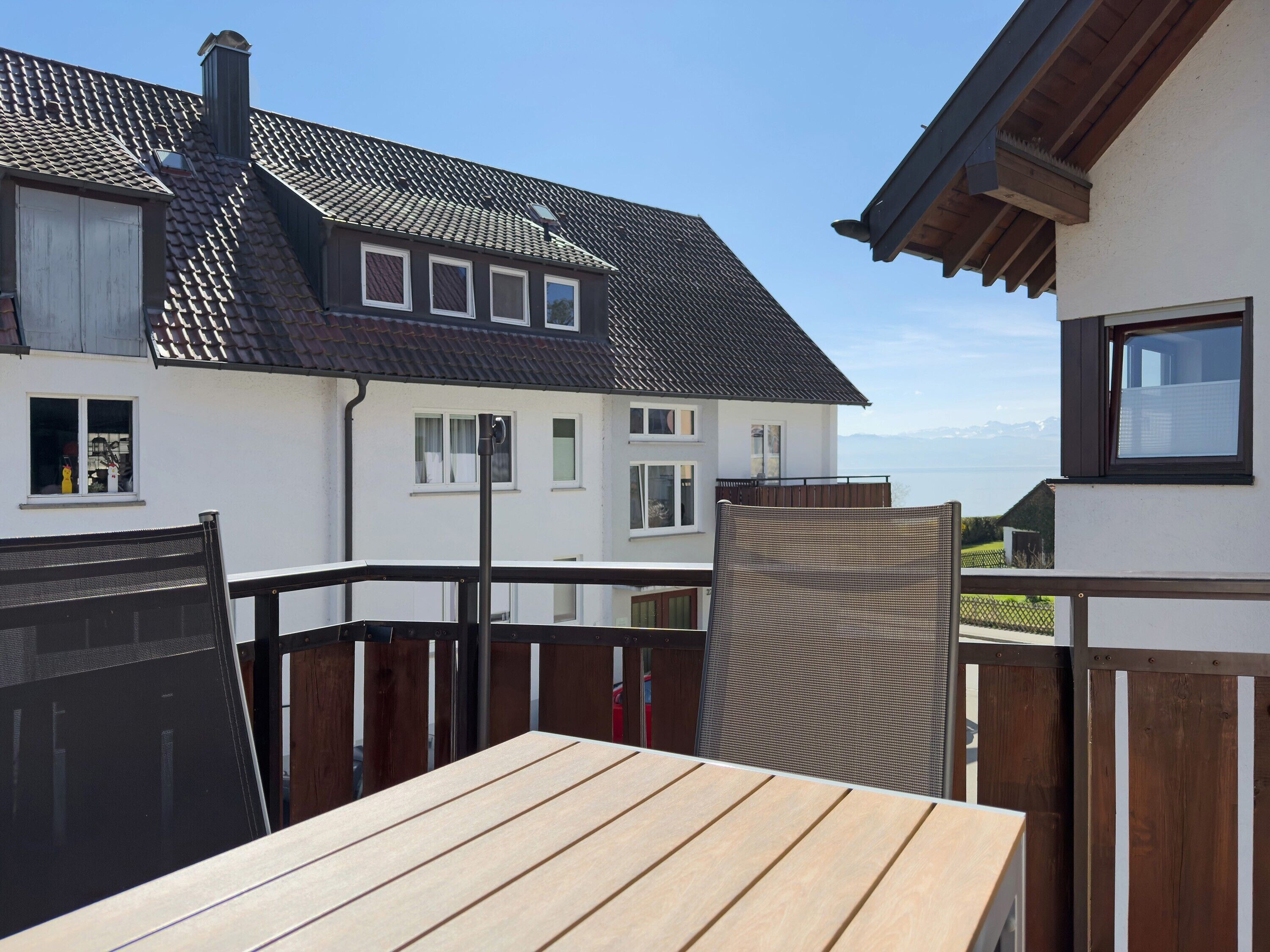 Une terrasse en bois avec une table, une chaise en métal et une vue sur une maison avec un toit en tuiles et de nombreuses fenêtres.