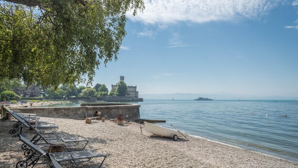 A pebbled beach with lounge chairs, a boat, and a historic building in the background.