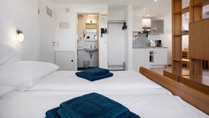 A hotel room with a bed, a bathroom, a kitchenette, and a wooden shelf unit. - Attic Apartment DG3 in "Gästehaus Heinemann" Directly on Lake Constance (Langenargen)