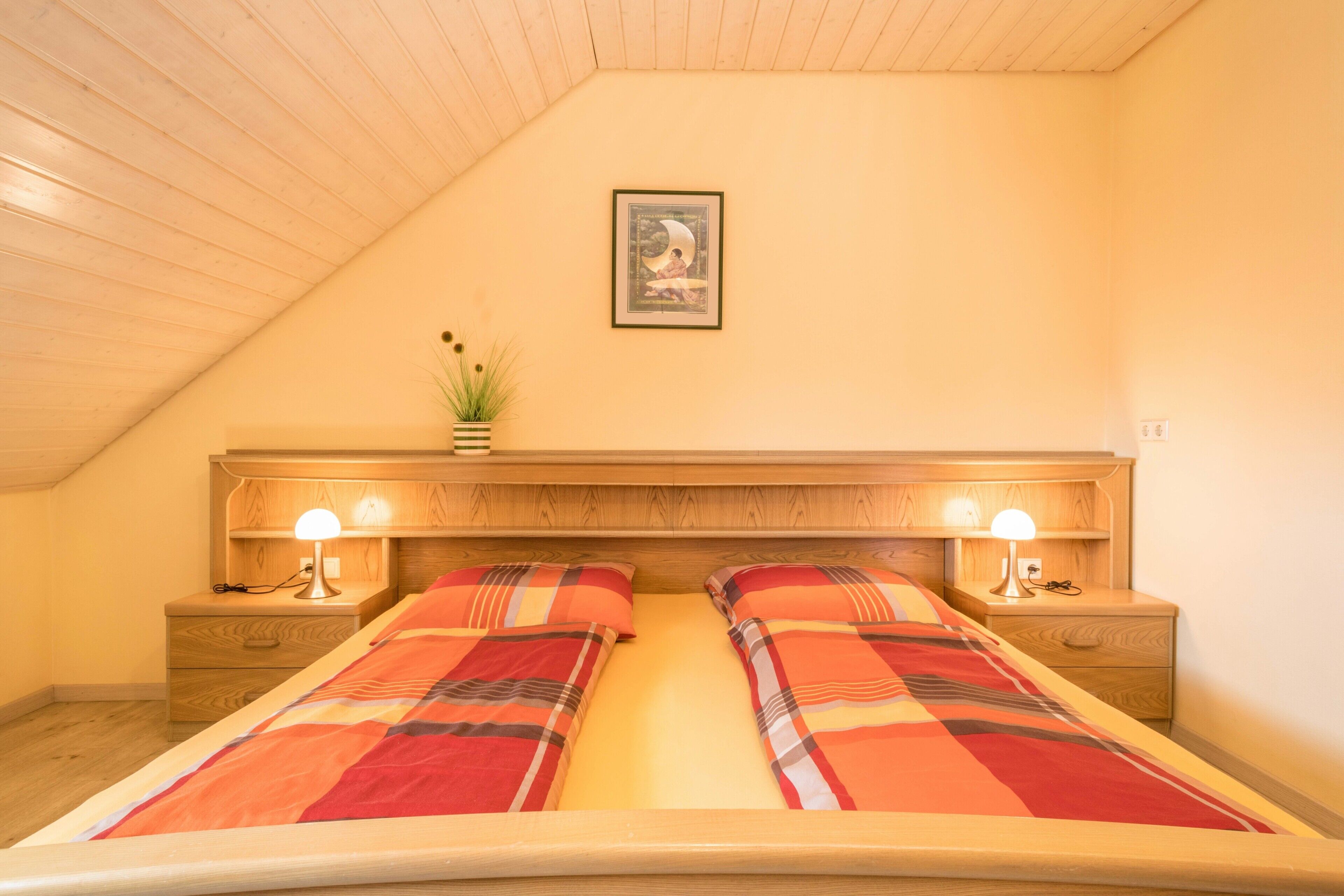 Ein Schlafzimmer mit Holzboden, einem Bett mit Karo-Bettwäsche, zwei Nachttischlampen, einem Nachttisch und einem gerahmten Bild an der Wand.