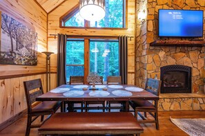 A wooden interior with a stone fireplace, a dining table with placemats, and a television mounted on the wall. - Pet-Friendly Broken Bow Cabin on Stream with Hot Tub, Dog Run & Game Room (Broken Bow)