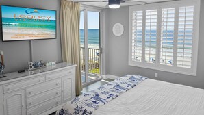 Un dormitorio con cama, cómoda, televisor mostrando 'Legacy Vacations', balcón con vista al océano y un cartel que dice 'Let the sand...'.