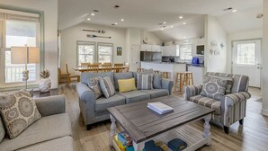 Ein großzügiges Wohnzimmer mit grauem Sofa, einem Couchtisch und einem gestreiften Sessel. Es gibt einen Essbereich mit Holzkohle und einem Tisch. Die Küche ist im Hintergrund mit weißen Schränken und einem Edelstahl-Kühlschrank zu sehen.