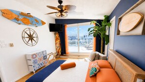 Un dormitorio con cama, ventilador de techo, ventana grande, planta y televisor instalado en la pared.