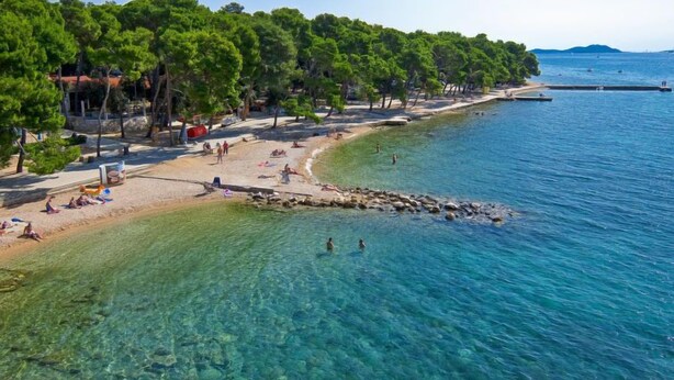 A beach with clear waters, people swimming and sunbathing, and a forested area in the background. - Apartment Petra by Interhome (Biograd na Moru)