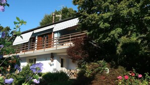 A two-story house with a balcony, surrounded by lush greenery and flowering plants.