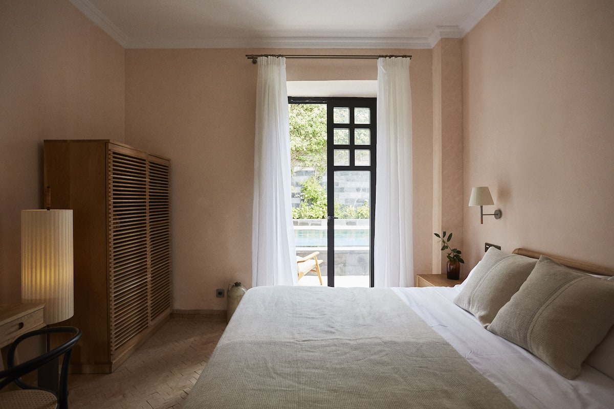 Une chambre avec un lit, une armoire en bois, une table de chevet avec une lampe, et une vue sur un espace extérieur, visible à travers une porte.