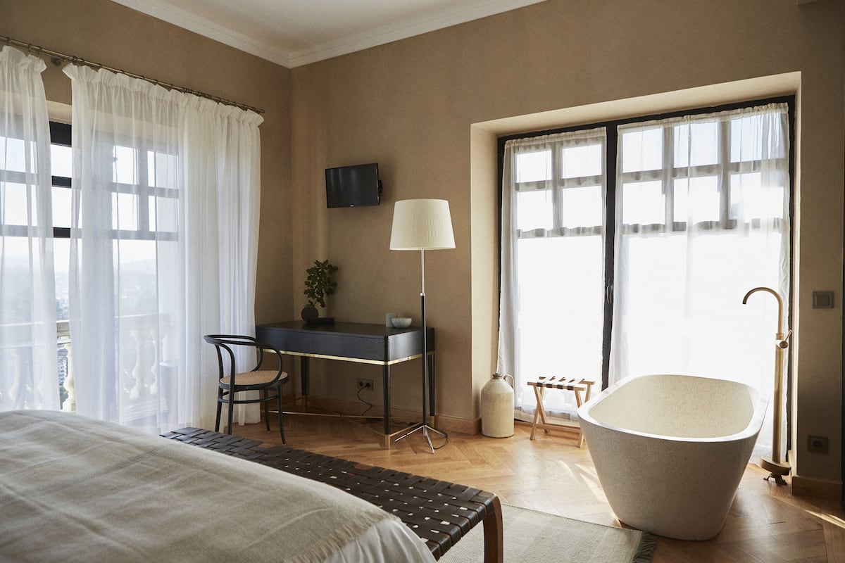 Une chambre d’hôtel comprenant un lit, un bureau avec une chaise, une grande baignoire, un téléviseur fixé au mur et une fenêtre avec des rideaux légers.