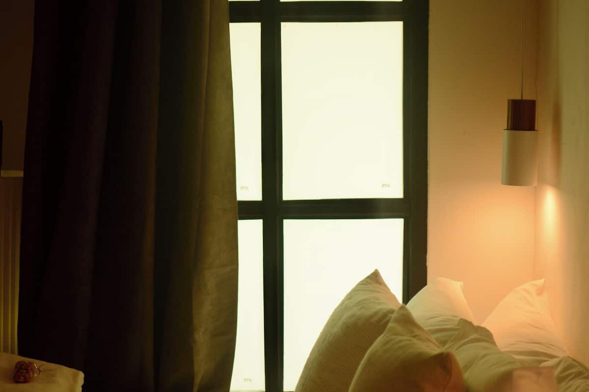 Un lit avec une literie blanche et plusieurs oreillers, une fenêtre donnant sur un beau paysage et un luminaire fixé au mur.
