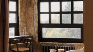 A rustic room with a stone wall, a large window, a wooden chair, a table with bottles, and a bathtub.