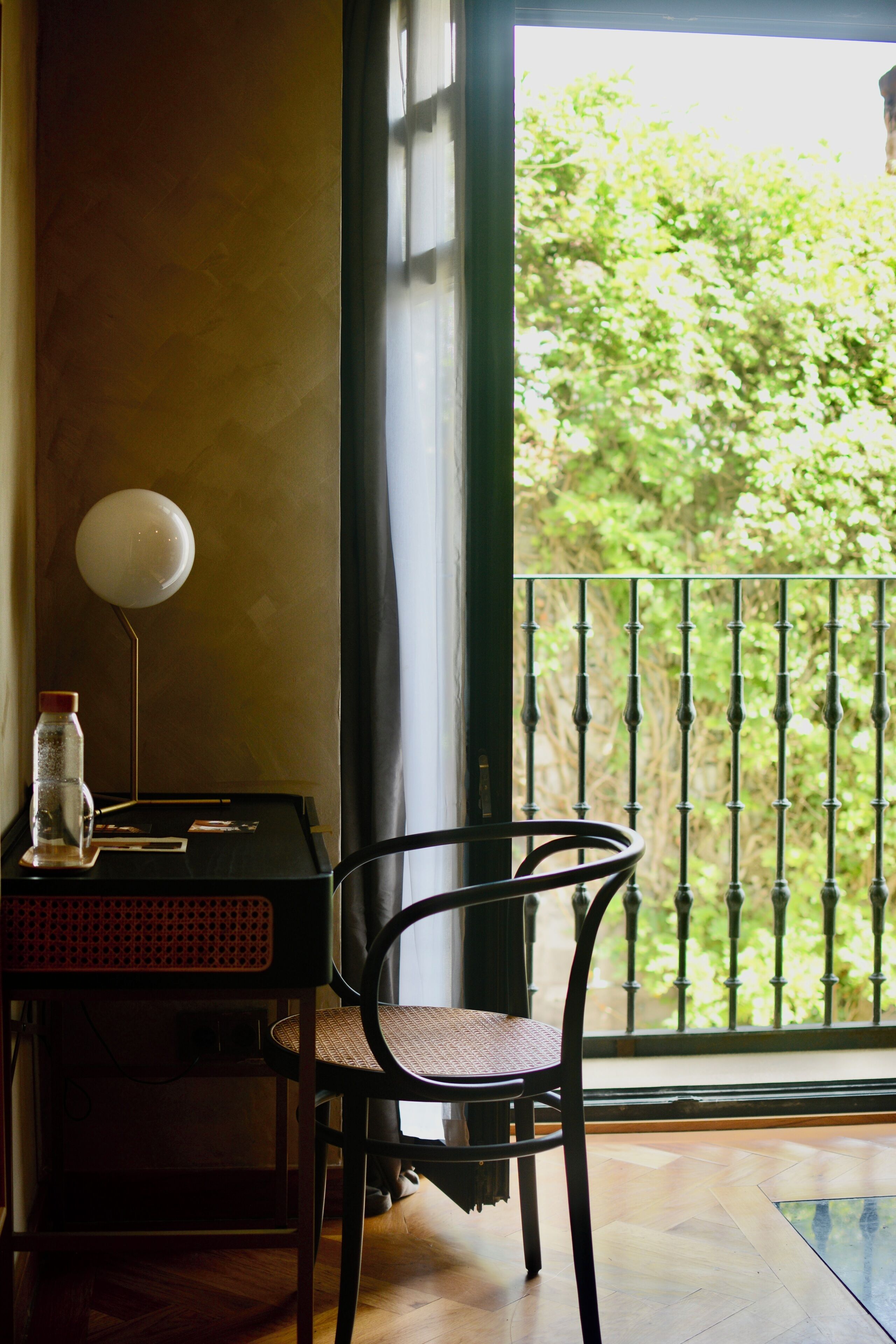 A room with a wooden floor, a black chair, a small table with a bottle and a lamp, and a view of greenery through a window with a black metal railing.