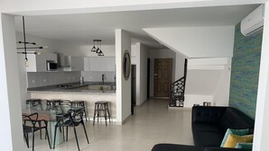 Una sala de estar moderna con un sofá negro y una mesa de comedor de vidrio con sillas. Se ve una cocina al fondo, con armarios blancos y una isla central.
