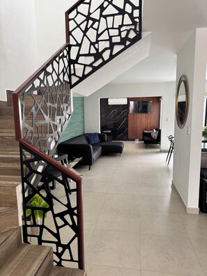 Una sala de estar moderna con un sofá seccional negro, una escalera de madera con barandilla geométrica y un espejo grande en la pared.