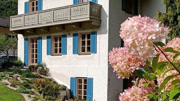 Ett hus i två våningar med en balkong på övervåningen, en trädgård med blommor och en klar, blå himmel.