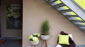 A staircase with a metal handrail, a bright yellow wall, a welcome mat, two potted plants, a patterned rug, and a white chair with a green pillow. - Ferienwohnung Riesling - Weinlodge am Geissberg (Eberstadt)