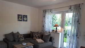 A modern living room with a grey sofa, patterned cushions, a small wooden table, and a glass coffee table. There are two motivational wall art pieces and a window with patterned curtains.
