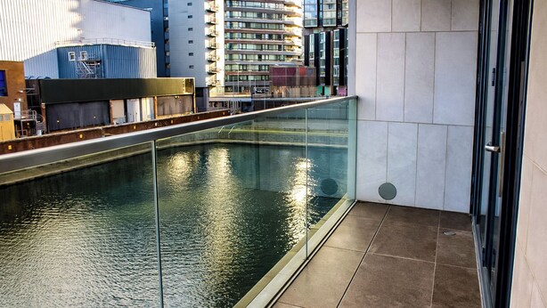 Un edificio moderno con un balcón de vidrio que da a una masa de agua, con edificios urbanos al fondo.