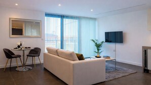 Una sala de estar moderna con un sofá, dos sillones, una mesita con una botella y vasos, un televisor y un ventanal grande con persianas.