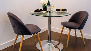 Un comedor con una mesa de vidrio, dos sillas, un jarrón con flores, una botella de vino y una copa de vino blanco.
