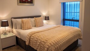Una cama bien hecha con una manta color beige, dos lámparas de noche y un cuadro en la pared.