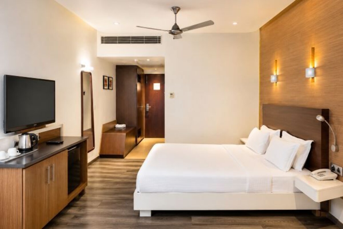 Una habitación de hotel con cama, televisor, escritorio, ventilador de techo e iluminación en la pared.