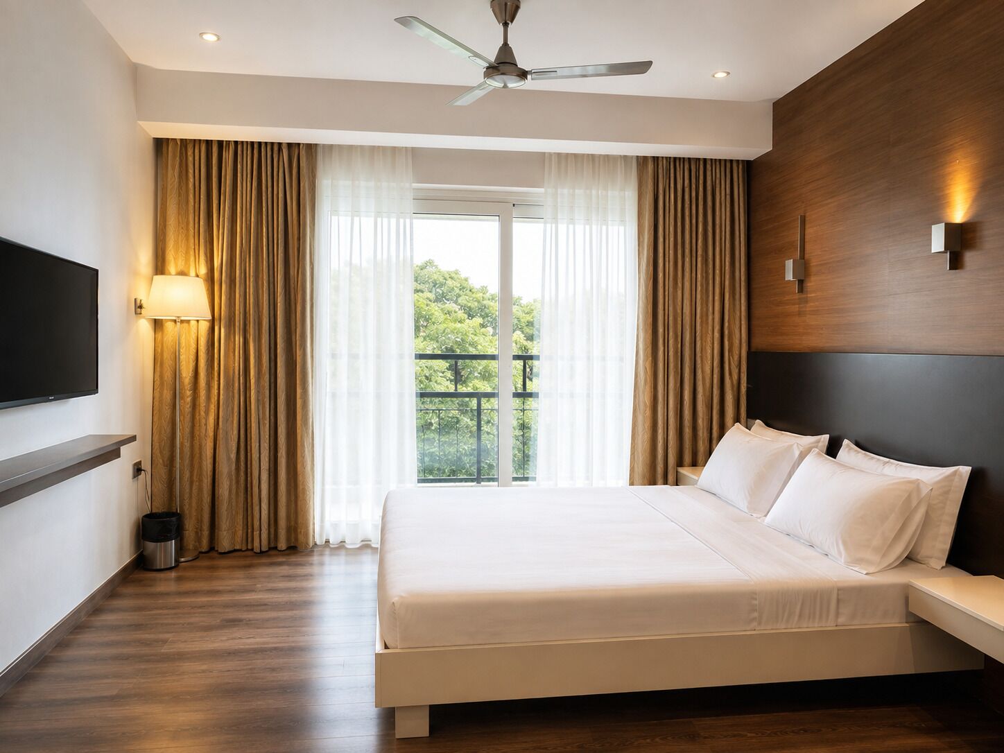 A hotel room with a bed, a TV, a ceiling fan, and a view of greenery outside.