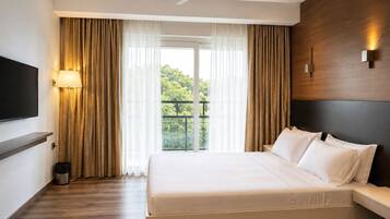 A hotel room with a bed, a TV, a ceiling fan, and a view of greenery outside.